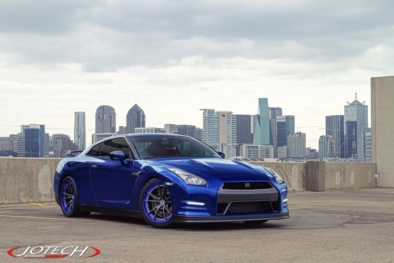 Blue Nissan GT-R with aftermarket wheels parked on a rooftop with a city skyline in the background.