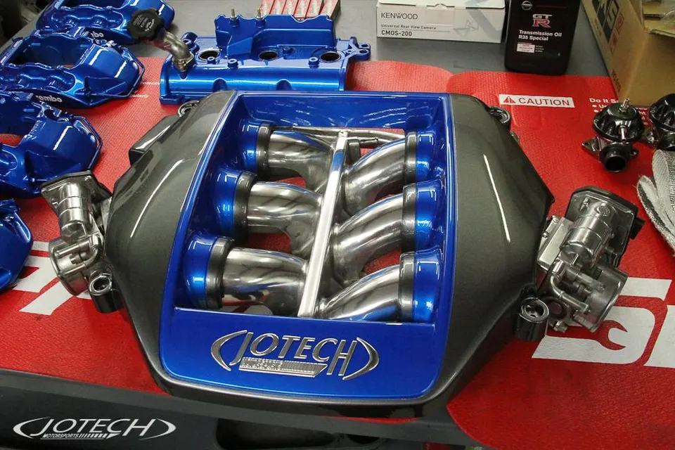 A Jotech Motorsports custom blue and grey intake manifold with individual throttle bodies and other engine parts on a workbench.