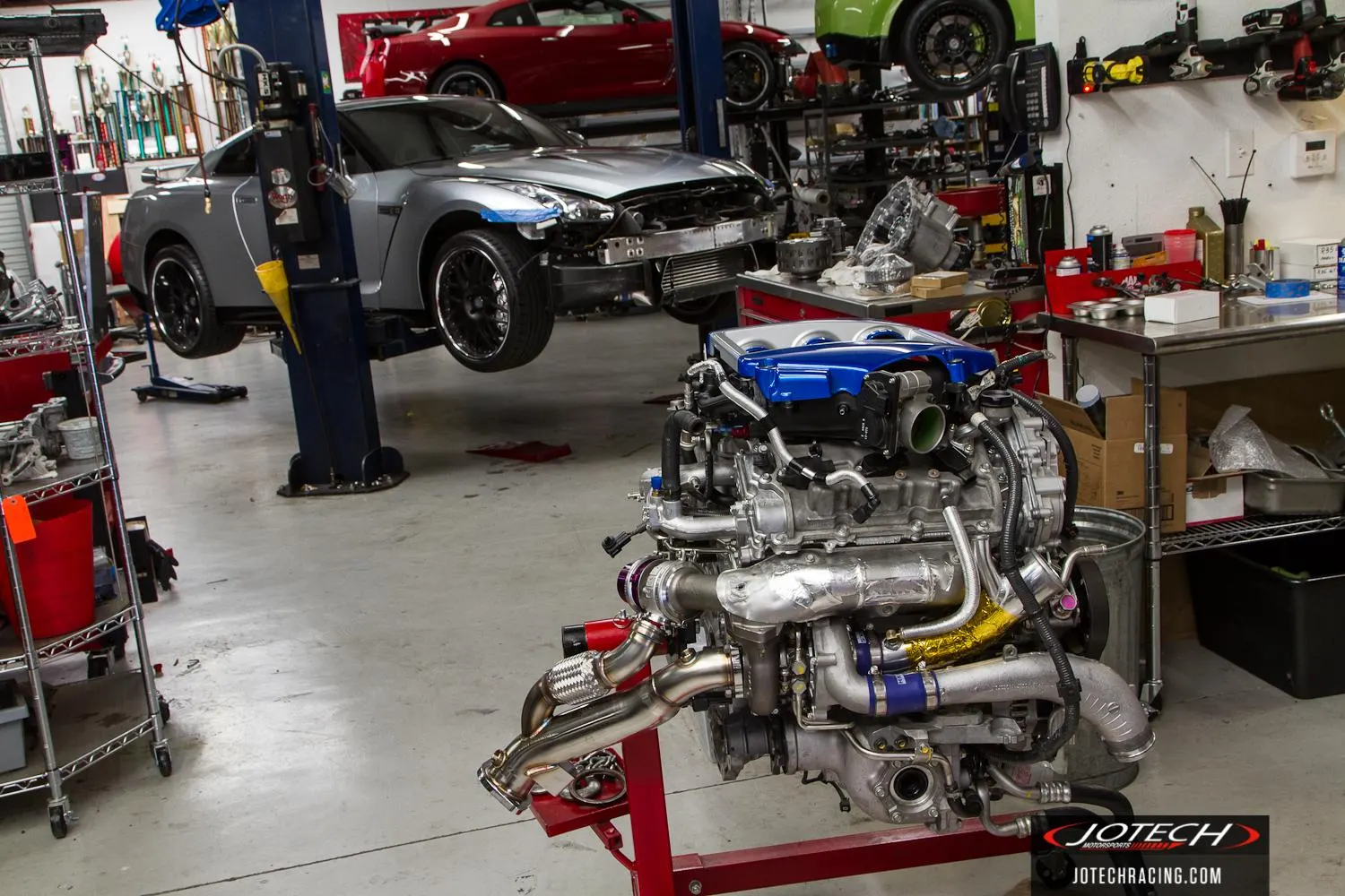 A Jotech Motorsports shop scene with a silver Nissan GT-R on a lift and a built VR38DETT engine on a stand.