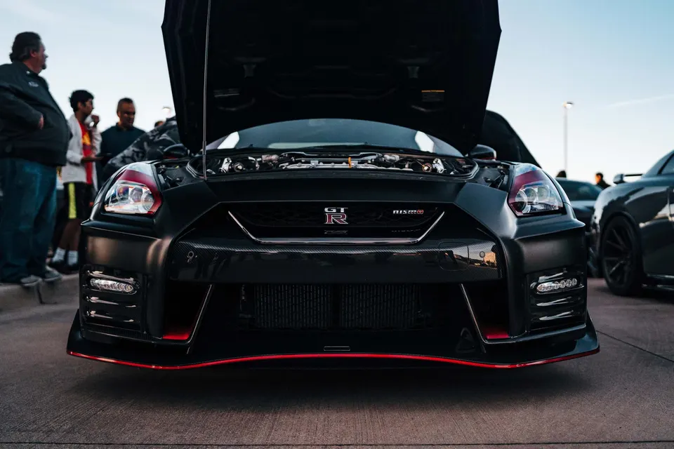 A black Nissan GT-R R35 with its hood open, featuring a custom front bumper with red accents and carbon fiber elements at a car event.