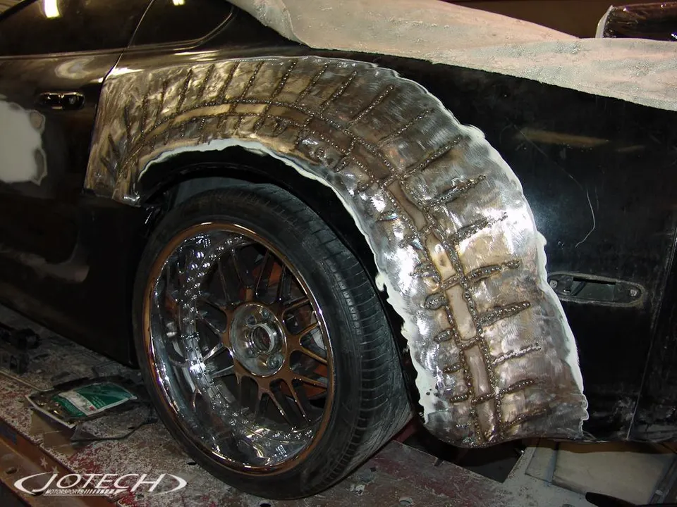 Custom widebody fender fabrication on a black car in a Jotech Motorsports shop, showing welding and chrome wheels.