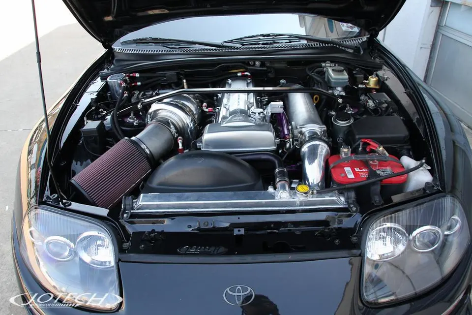 Engine bay of a black Toyota Supra with a large aftermarket turbocharger and intake system, showcasing performance modifications.