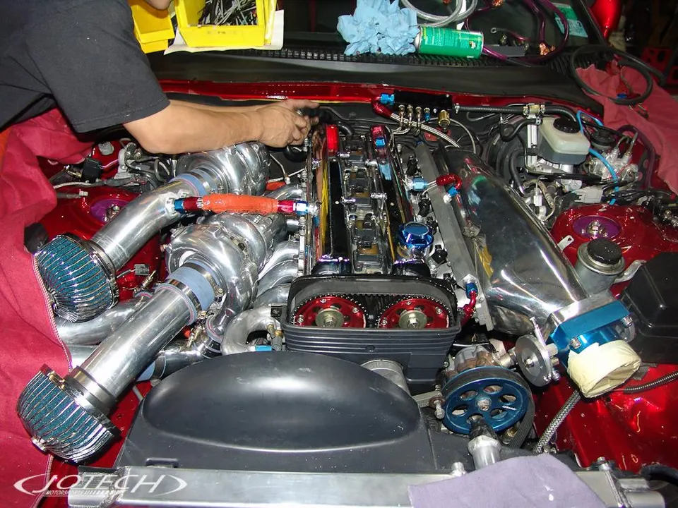 A mechanic works on a highly modified twin-turbo engine in a red Toyota Supra, featuring custom intake and boost piping.