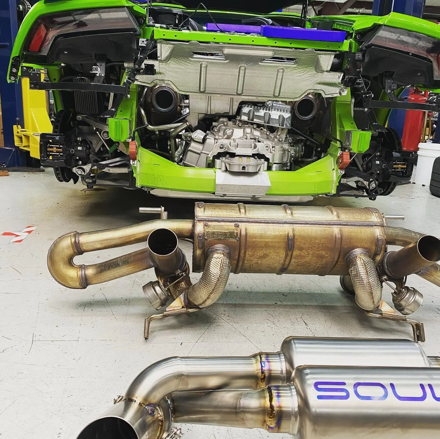 A green Lamborghini Huracan on a lift in a shop with its OEM exhaust removed and a new Soul Performance exhaust system ready for installation.