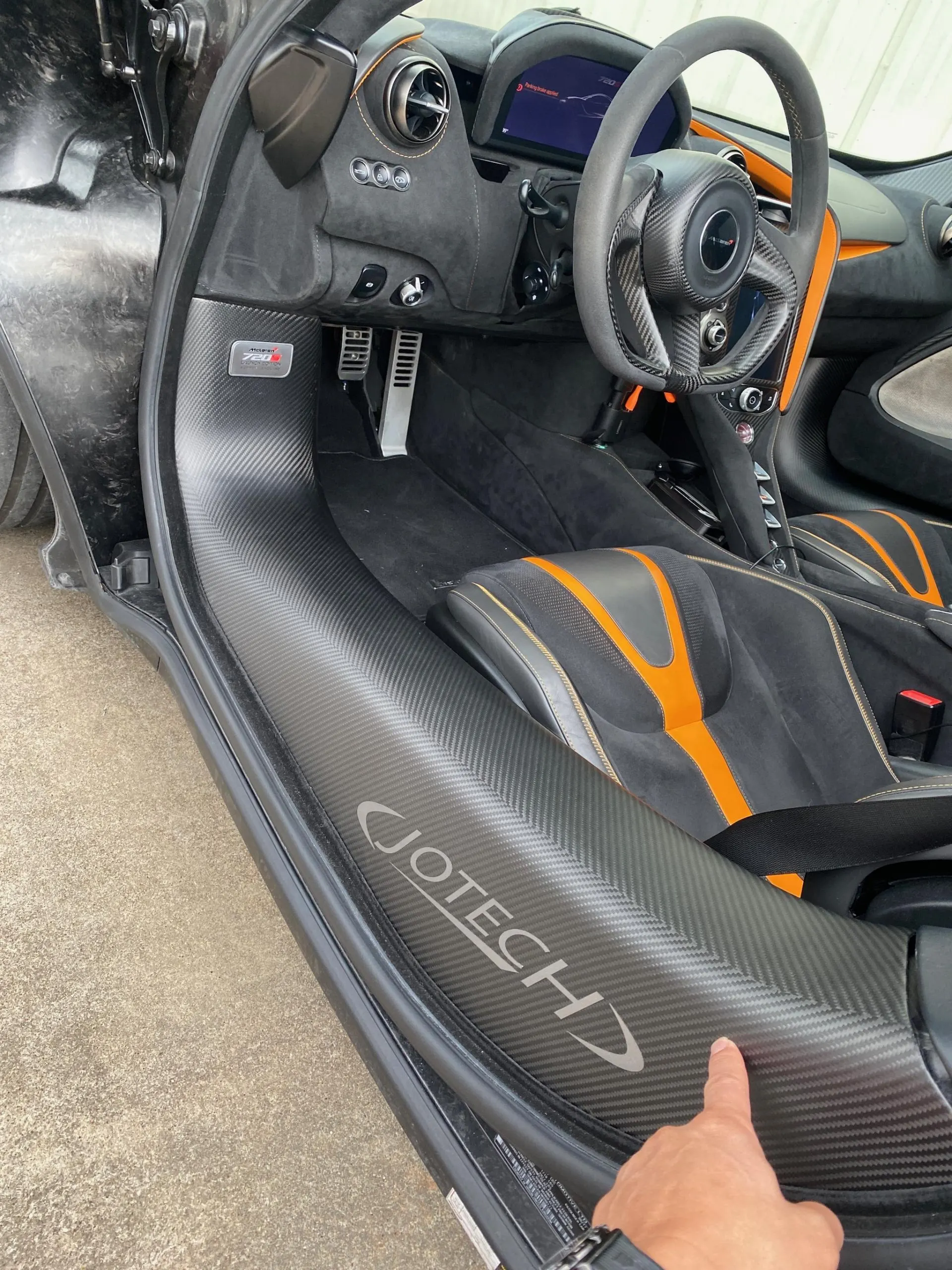 Close-up of a McLaren interior featuring carbon fiber door sills with Jotech branding, steering wheel, and orange accented seats.