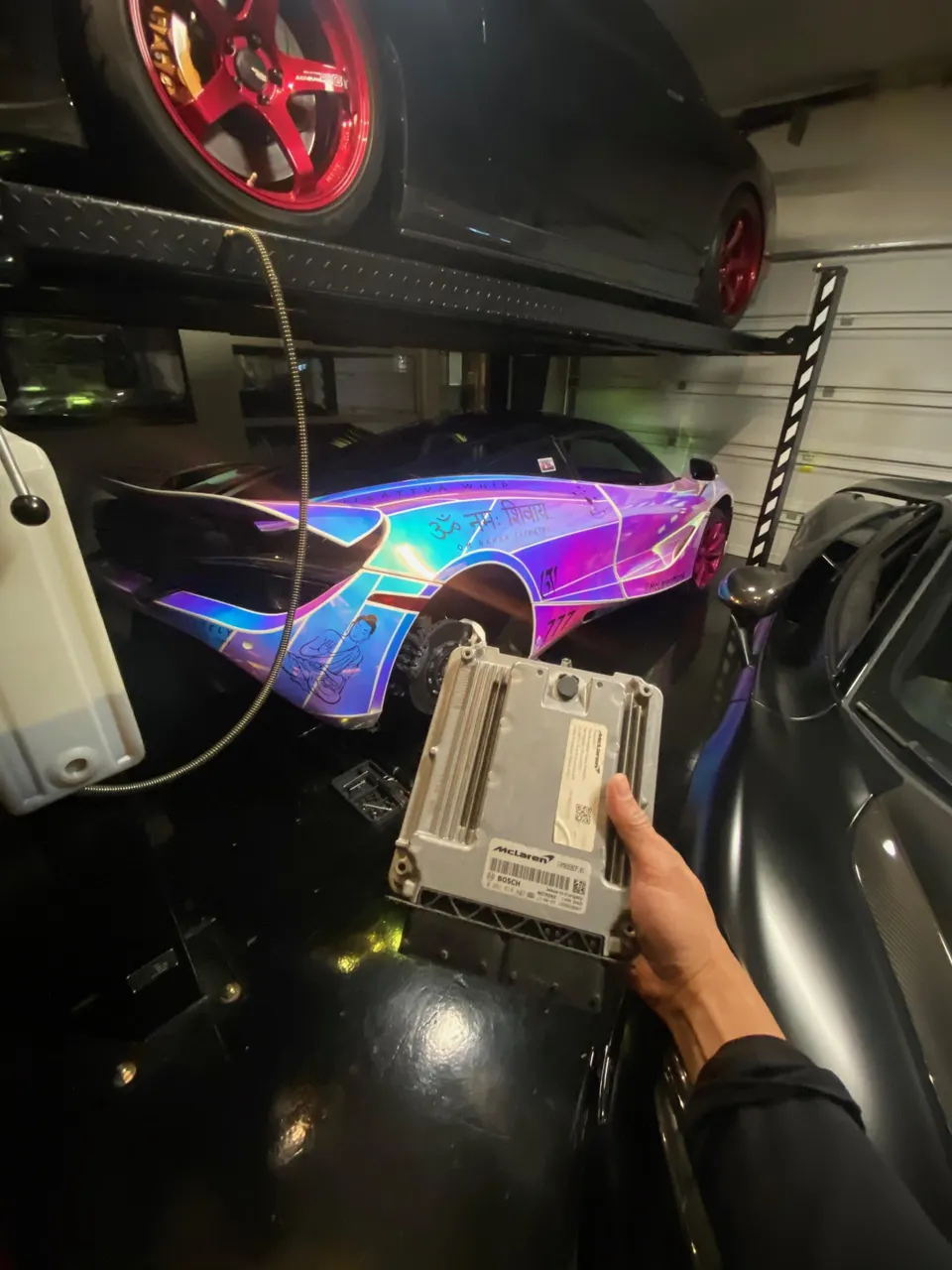 A hand holding a McLaren ECU in a performance tuning shop with multiple supercars, including a wrapped McLaren.