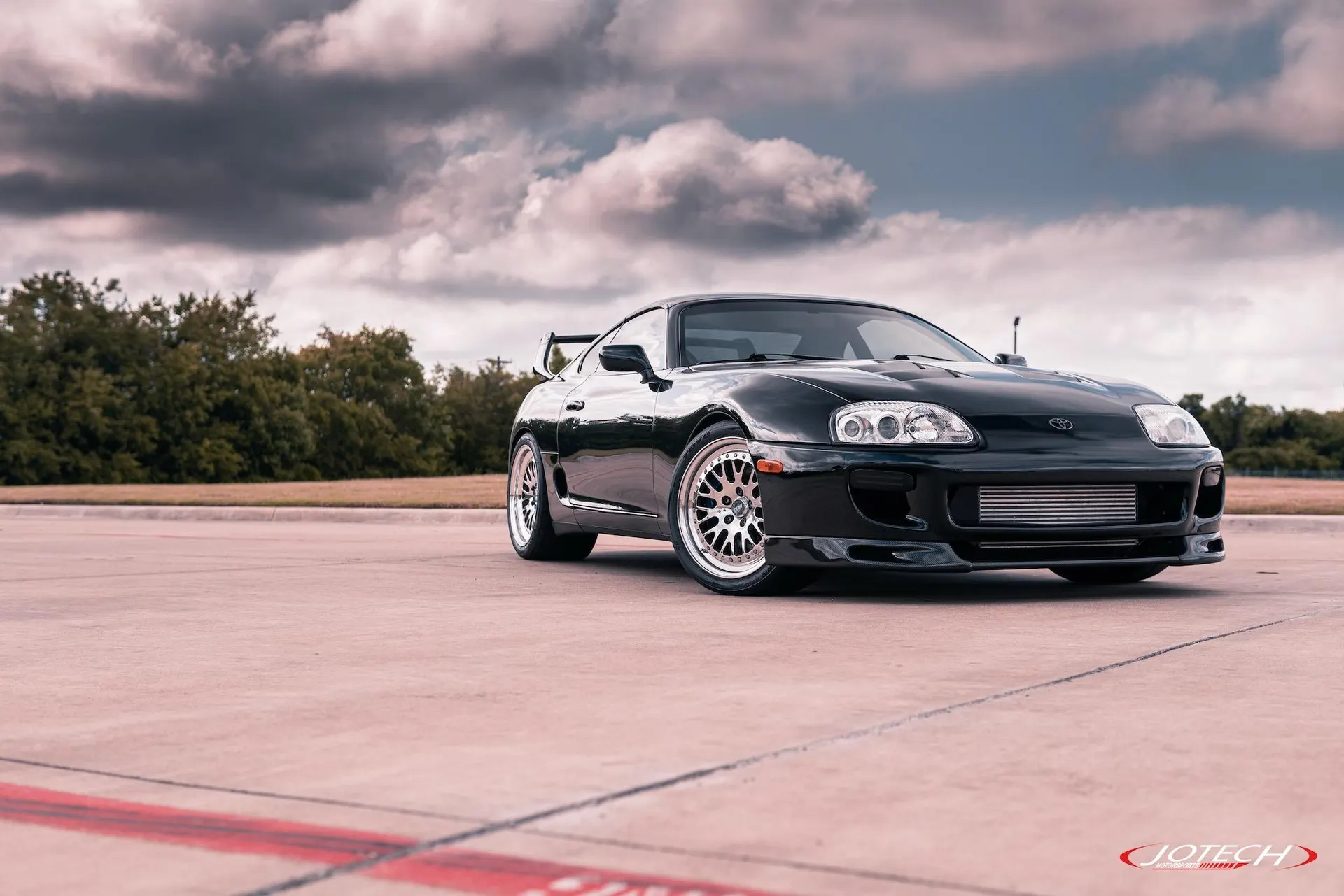 Black Toyota Supra MK4 with aftermarket wheels and a visible intercooler parked on an asphalt lot under a cloudy sky.