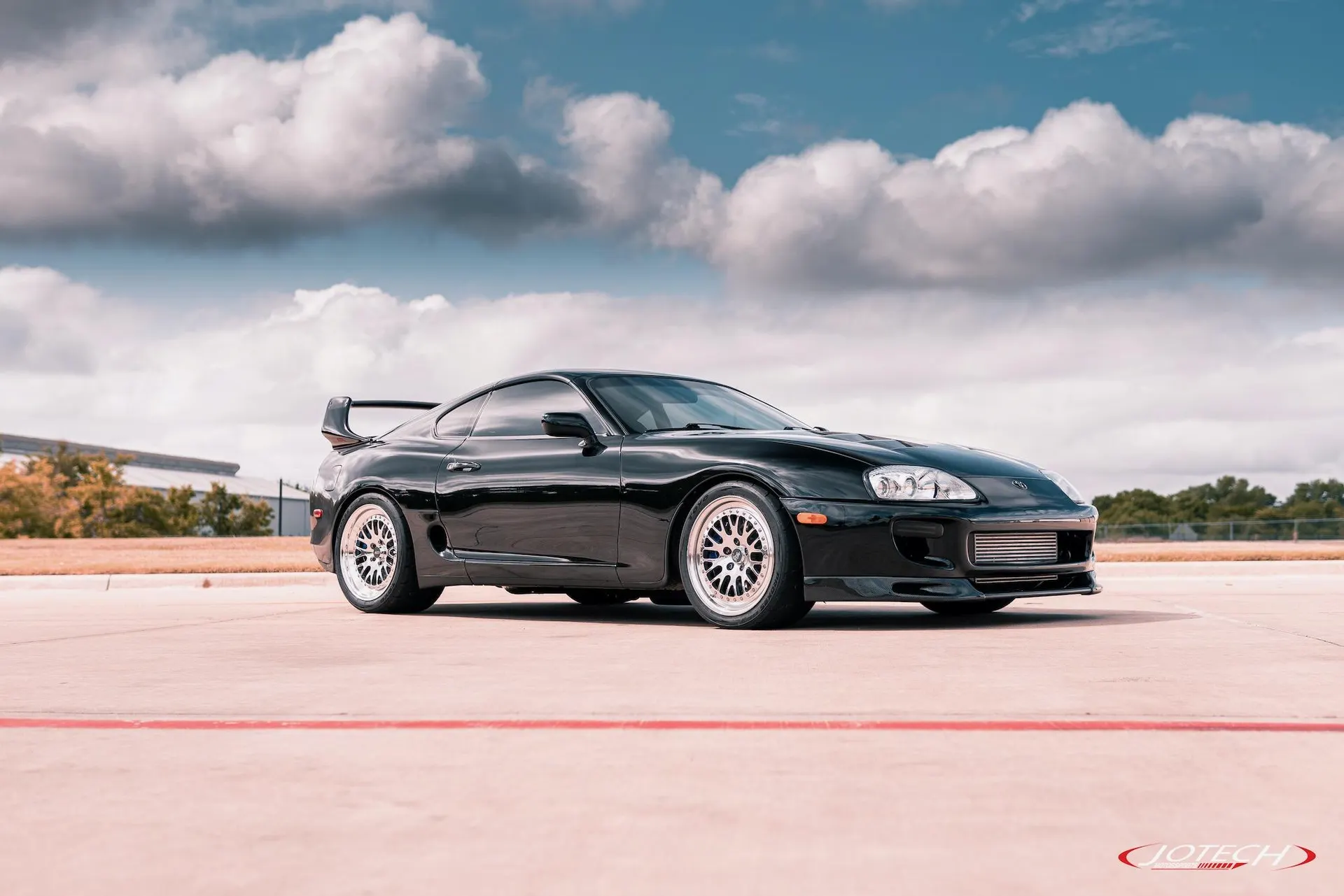 Black Toyota Supra MK4 with aftermarket wheels and a large front-mounted intercooler parked outdoors under a cloudy sky.