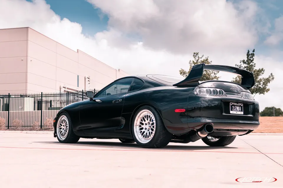 Black Toyota Supra MK4 with aftermarket multi-spoke wheels, large rear wing, and custom exhaust parked in front of a building.