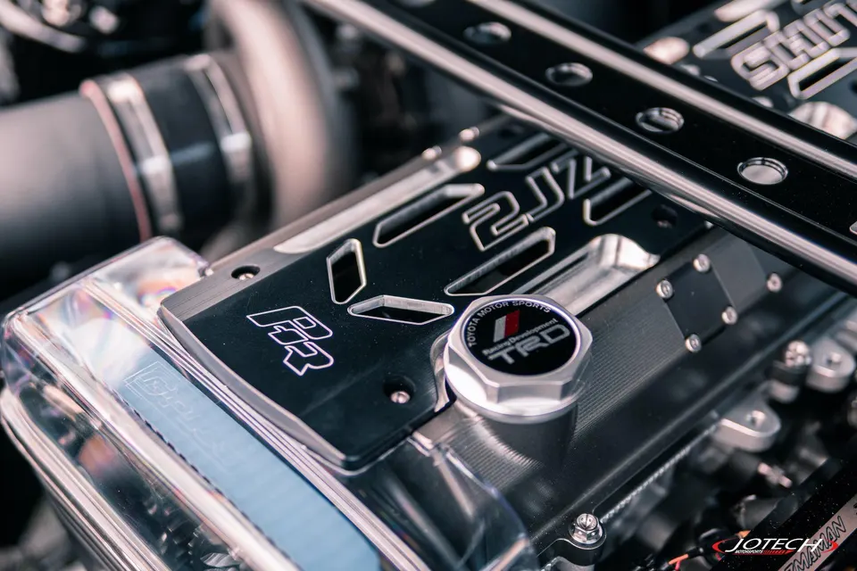 A close-up of a custom 2JZ engine bay with a black valve cover featuring '2JZ' and 'TRD' branding.