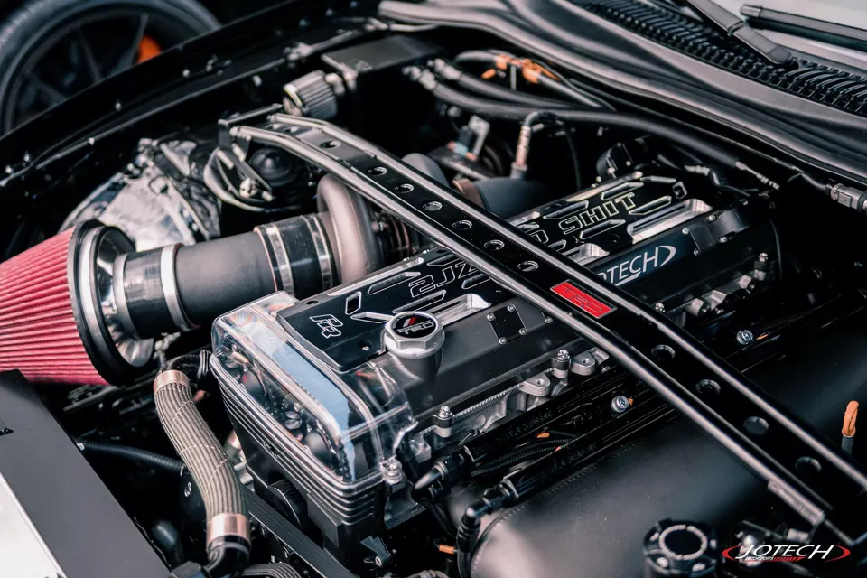 A close-up of a modified 2JZ engine bay featuring a large red air filter, turbocharger, and custom valve cover.