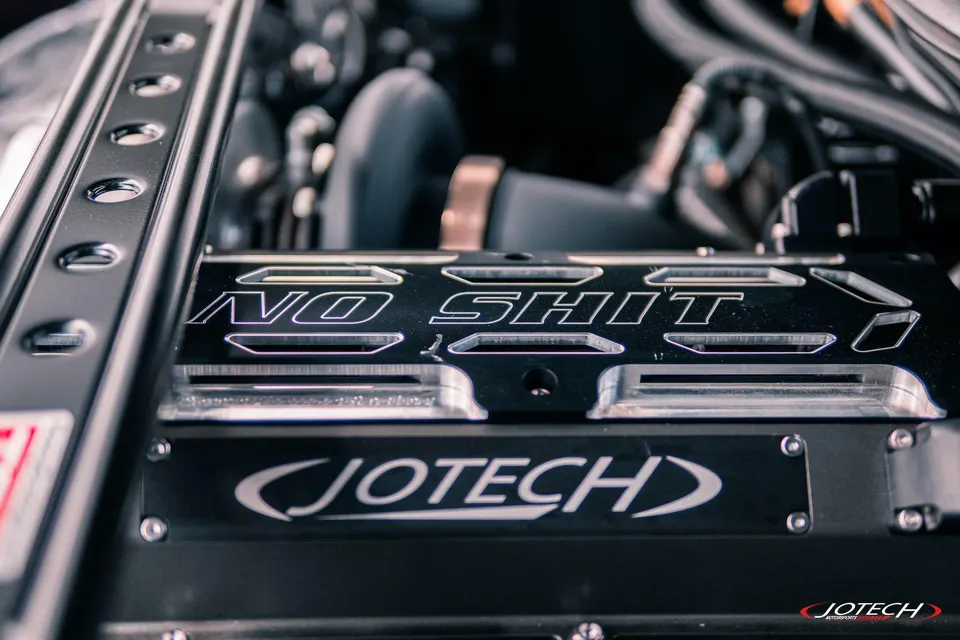 Close-up detail of a custom Jotech engine cover with 'NO SHIT' engraving in a performance car's engine bay.
