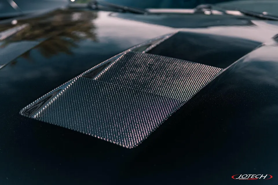 Close-up of a car's glossy black hood featuring a textured carbon fiber vent or scoop.