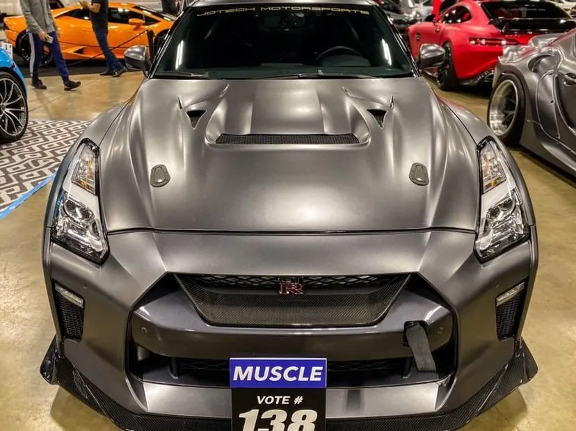 Front view of a matte grey Nissan GT-R with a carbon fiber splitter and hood pins at a car show.
