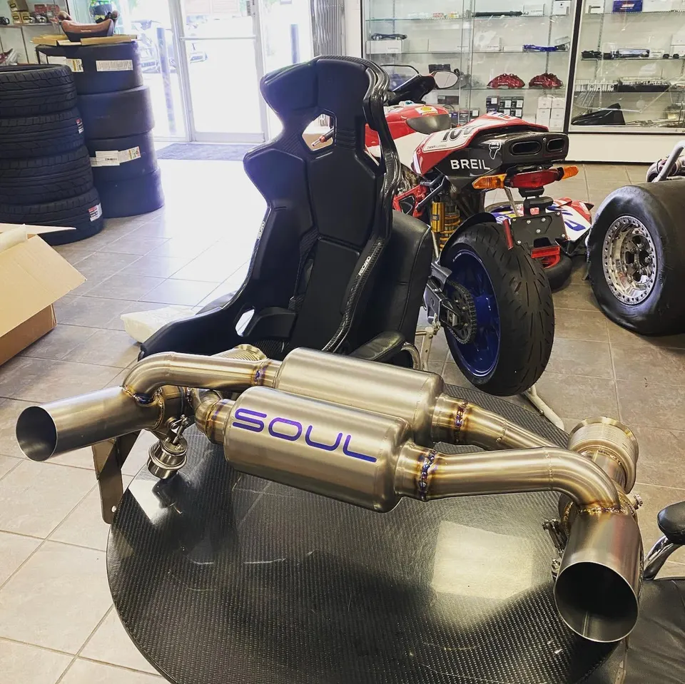 Soul Performance exhaust system displayed on a table in a performance tuning shop with a racing seat and motorcycle in the background.