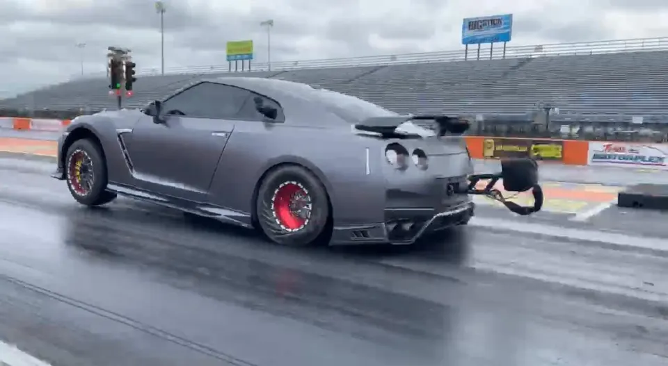 Gray Nissan GT-R with drag slicks and parachute launching on a wet drag strip with grandstands in the background.