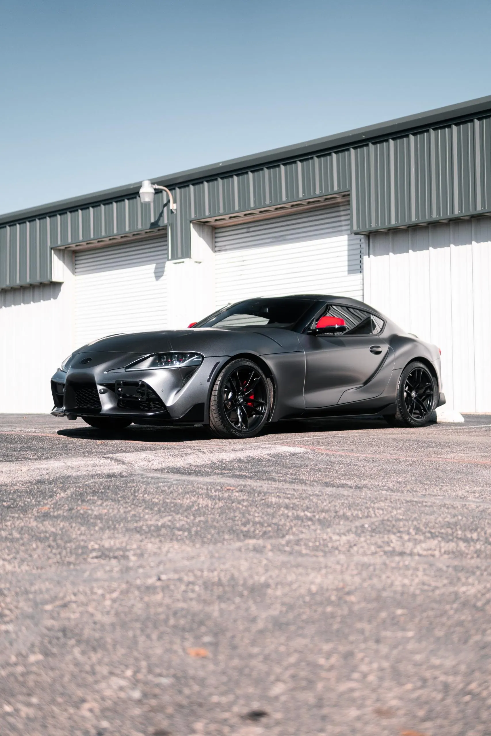 Matte grey Toyota Supra with red side mirrors and black wheels parked in front of a garage building.
