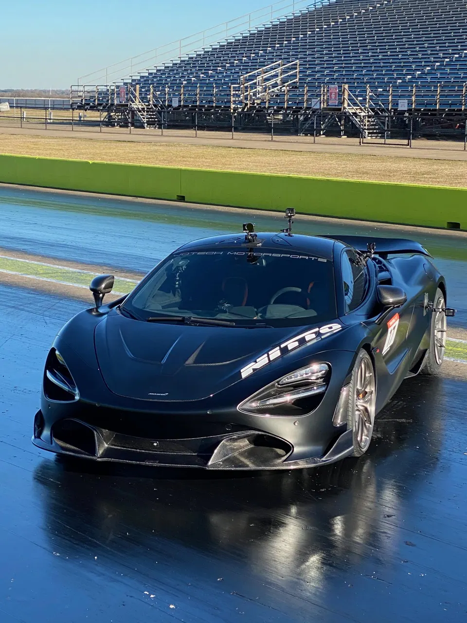 Black McLaren 720S supercar with carbon fiber aero on a drag strip with grandstands in the background, ready for a run.