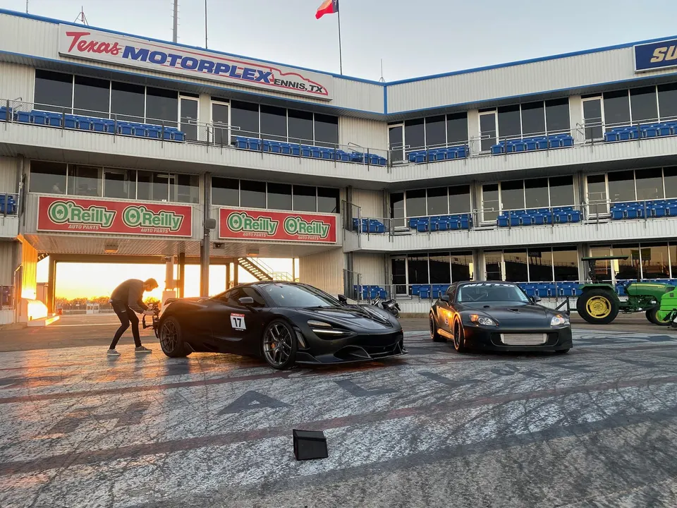 A McLaren and a Honda S2000 at the Texas Motorplex drag strip during sunset, with a person filming.