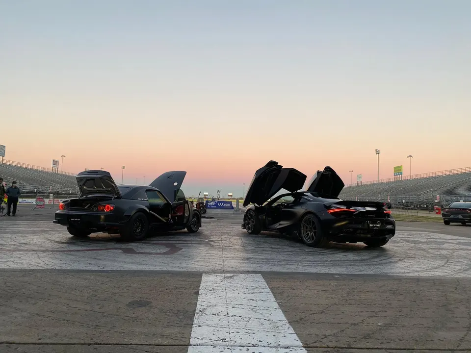 Two modified cars, a Mazda RX-8 and a McLaren 720S, with doors open at a drag strip during sunset.