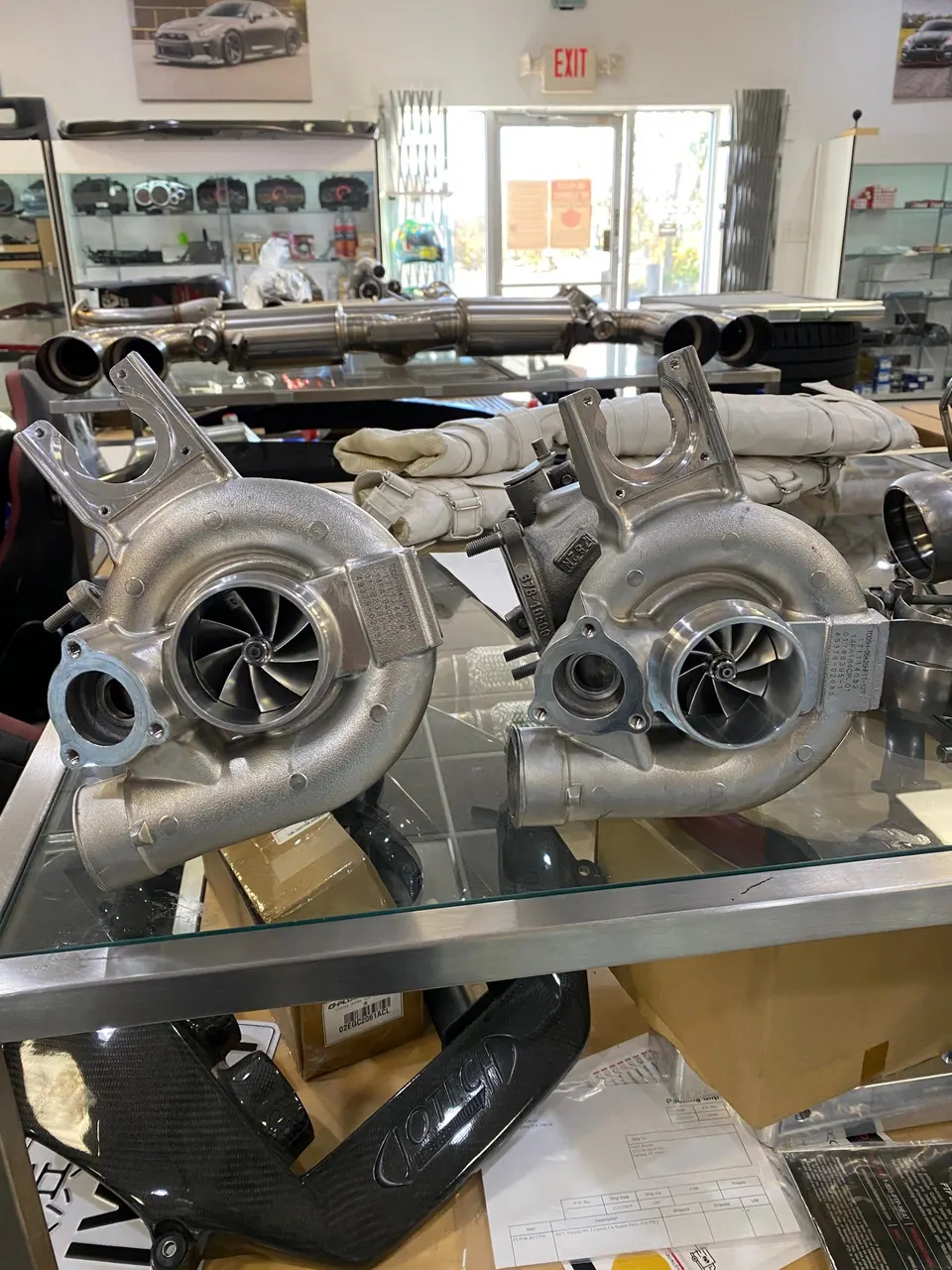 Two high-performance turbochargers on display in a tuning shop, with other aftermarket parts and an exhaust system in the background.