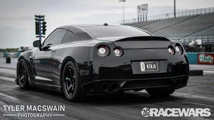 A black Nissan GT-R with a custom 'T NINJA' license plate on a drag strip, viewed from the rear with starting lights in the background.