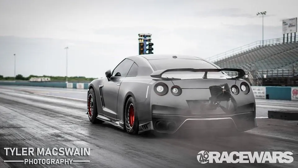 Grey Nissan GT-R with a large rear wing, parachute, and red wheels performing a burnout on a drag strip.