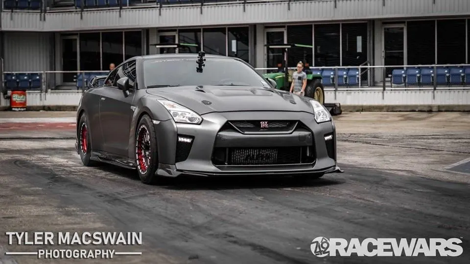 A grey Nissan GT-R on a drag strip with a building and bleachers in the background, ready for a race.