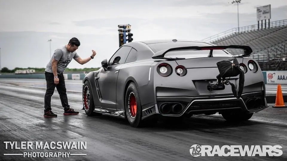Grey Nissan GT-R with a parachute and large rear wing on a drag strip, with a man standing next to the car.