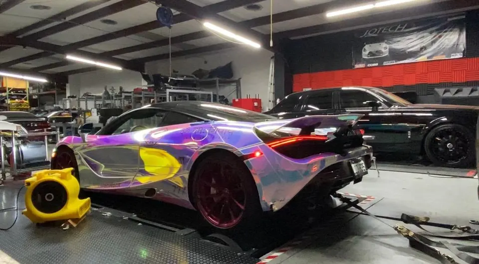 A McLaren 720S with an iridescent wrap on a dyno at a performance tuning shop, with a Rolls Royce in the background.