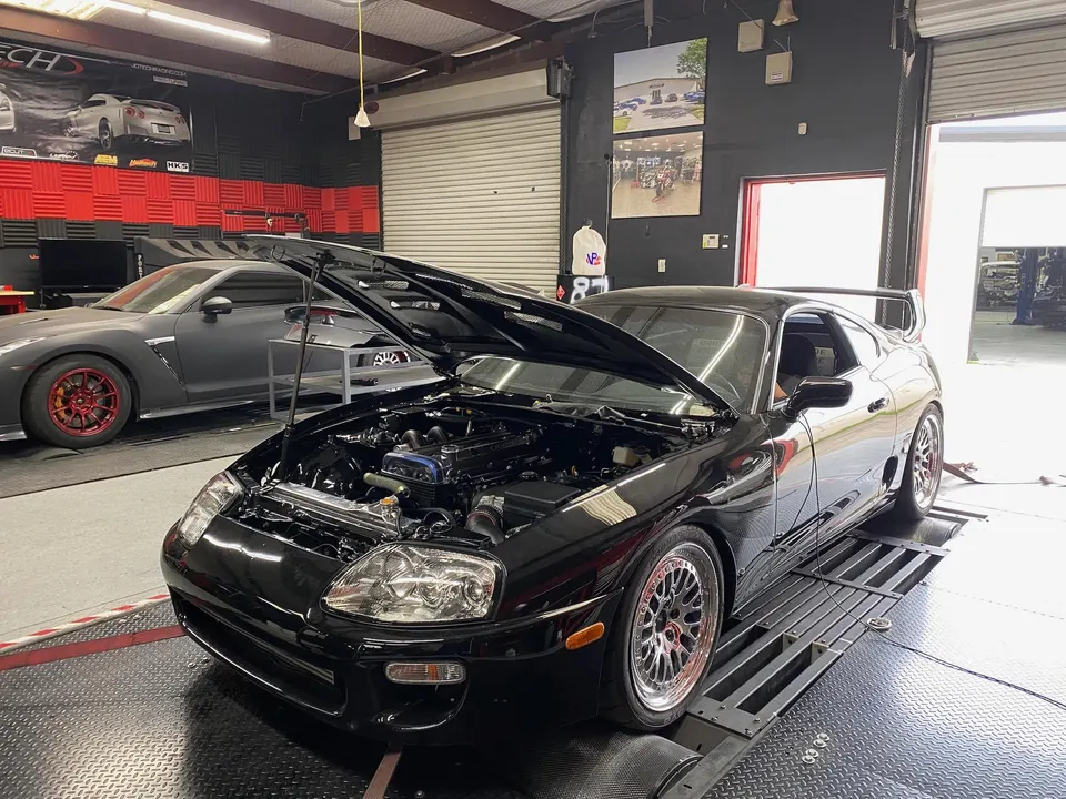 A black Toyota Supra with its hood open on a dyno at Jotech Motorsports, with a grey Nissan GT-R in the background.