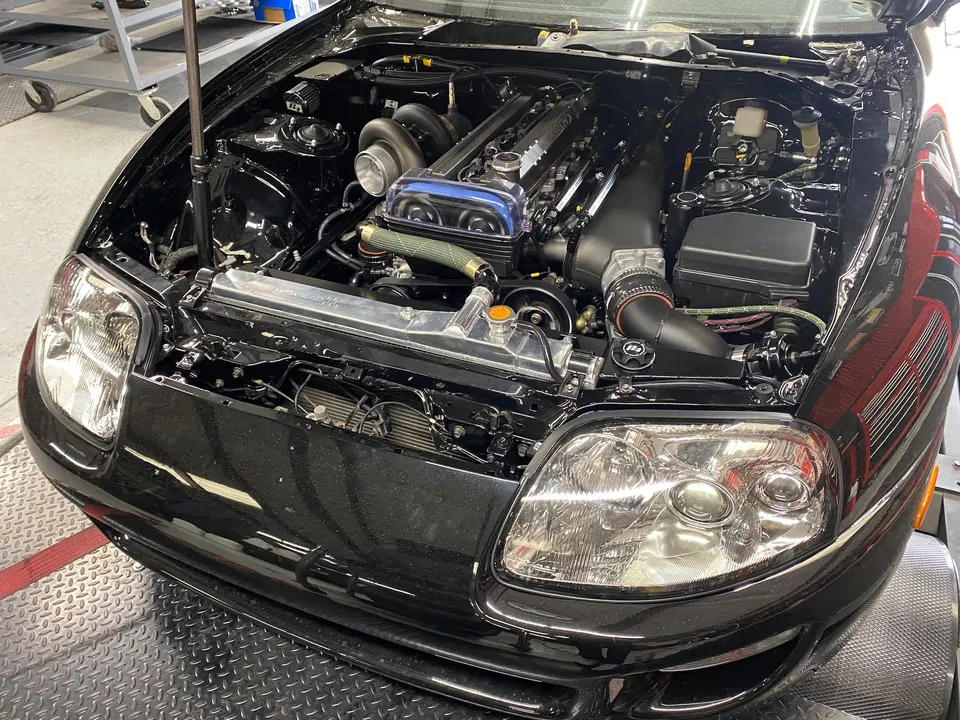 A black Toyota Supra with its hood open, revealing a highly modified 2JZ engine with a large turbocharger in a performance shop.