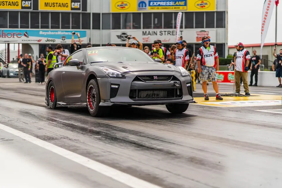 Grey Nissan GT-R with red wheels in the staging lane at a drag strip, surrounded by spectators.