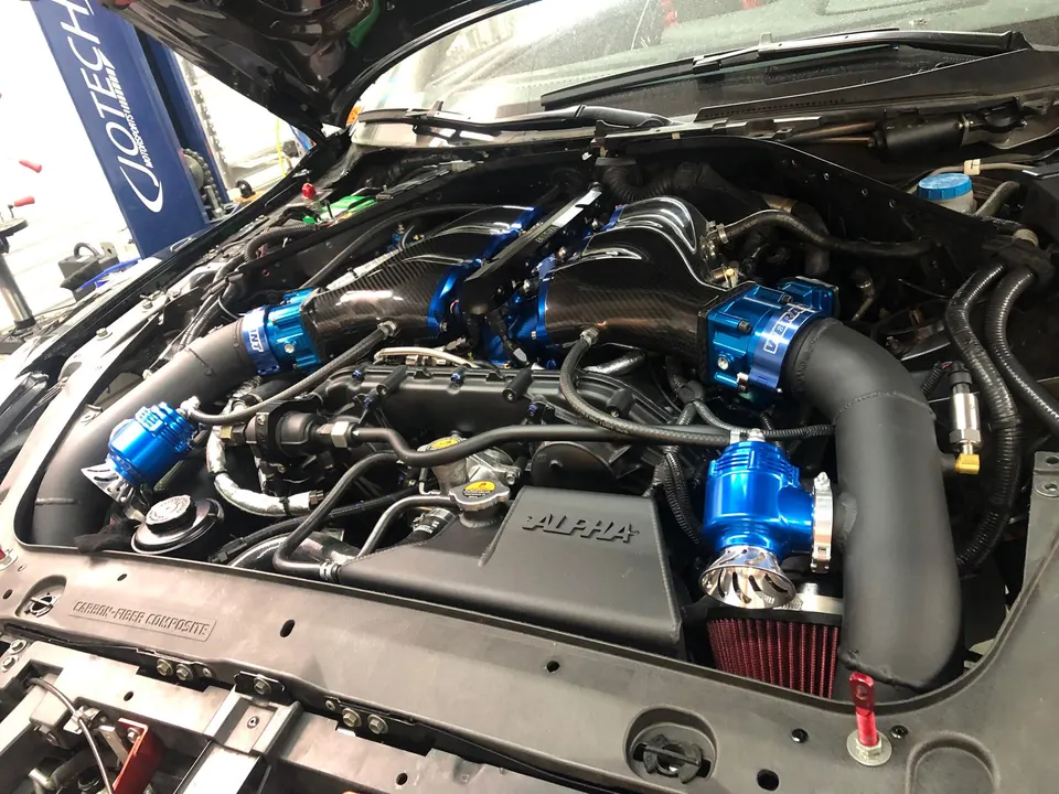 A detailed view of a high-performance car's engine bay featuring a carbon fiber intake manifold, blue blow-off valves, and aftermarket boost piping.