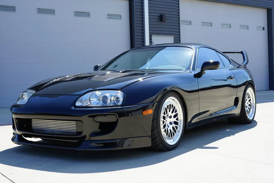 Black Toyota Supra MK4 with aftermarket wheels, carbon fiber hood, and large front-mount intercooler parked outside.