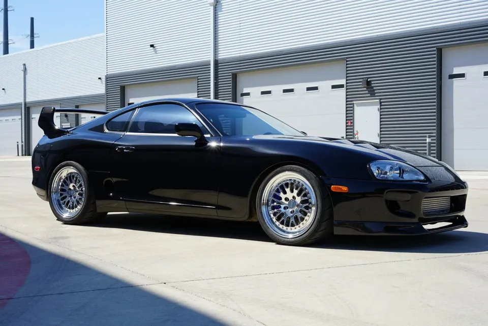 A black Toyota Supra MK4 with aftermarket wheels and a large rear wing parked in front of a modern building.