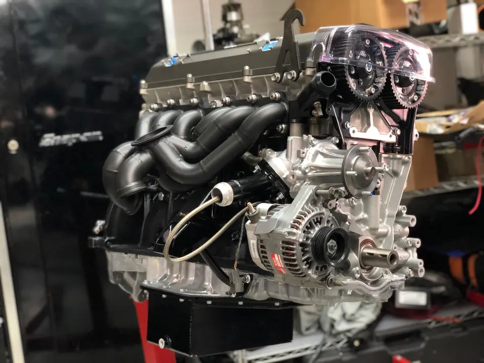 A detailed view of a high-performance 2JZ engine assembly with a black exhaust manifold and clear timing cover in a shop.