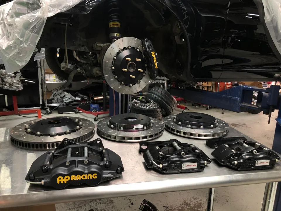 AP Racing brake rotors and calipers laid out on a table next to a car on a lift in a performance shop.