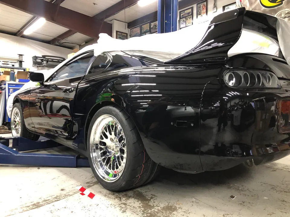 A black Toyota Supra on a lift in a shop, featuring aftermarket chrome wheels with drag tires and a large rear wing.