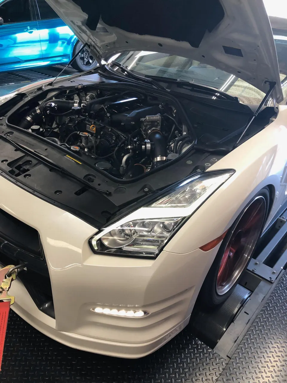 White Nissan GT-R with hood open on a dyno, showcasing the engine bay with performance induction and turbo piping.