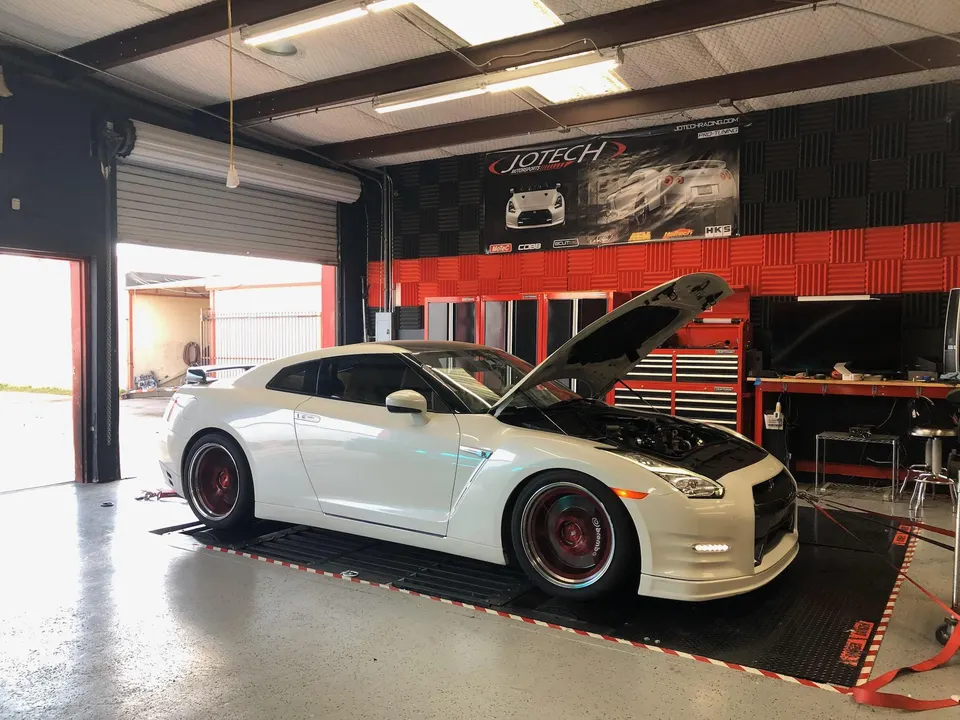 A white Nissan GT-R with its hood open is strapped to a dyno at Jotech Motorsports for performance tuning.