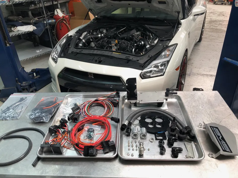 White Nissan GT-R with hood open in a shop, with aftermarket fuel system components and wiring harnesses laid out on a table.