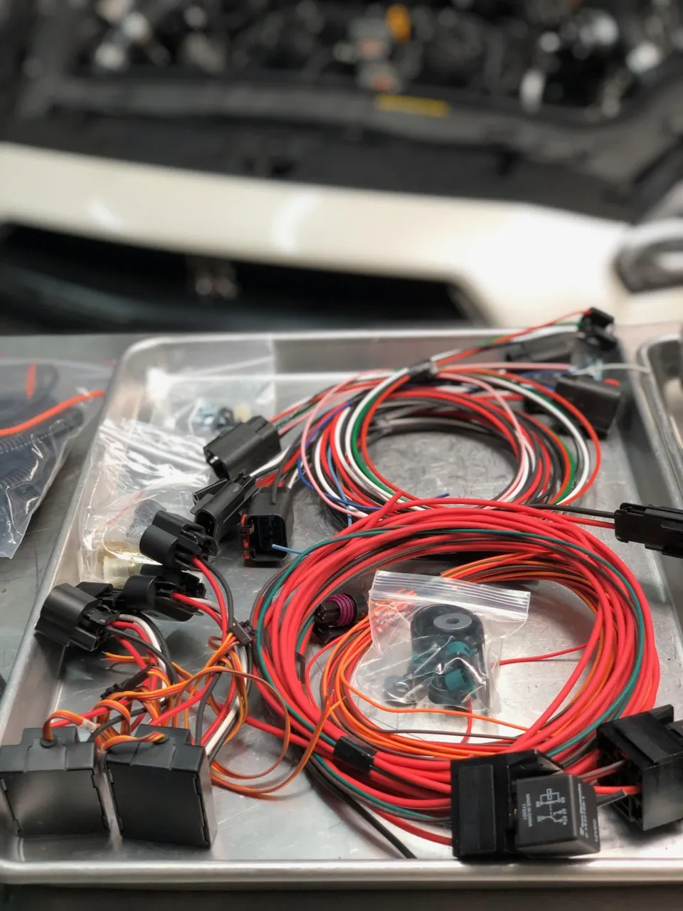 New wiring harness, electrical connectors, and relays laid out on a metal tray in a performance shop, with a car engine bay in the background.