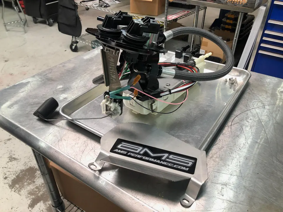 A detailed view of an AMS Performance fuel pump assembly with a fuel level sender on a metal workbench in a shop.