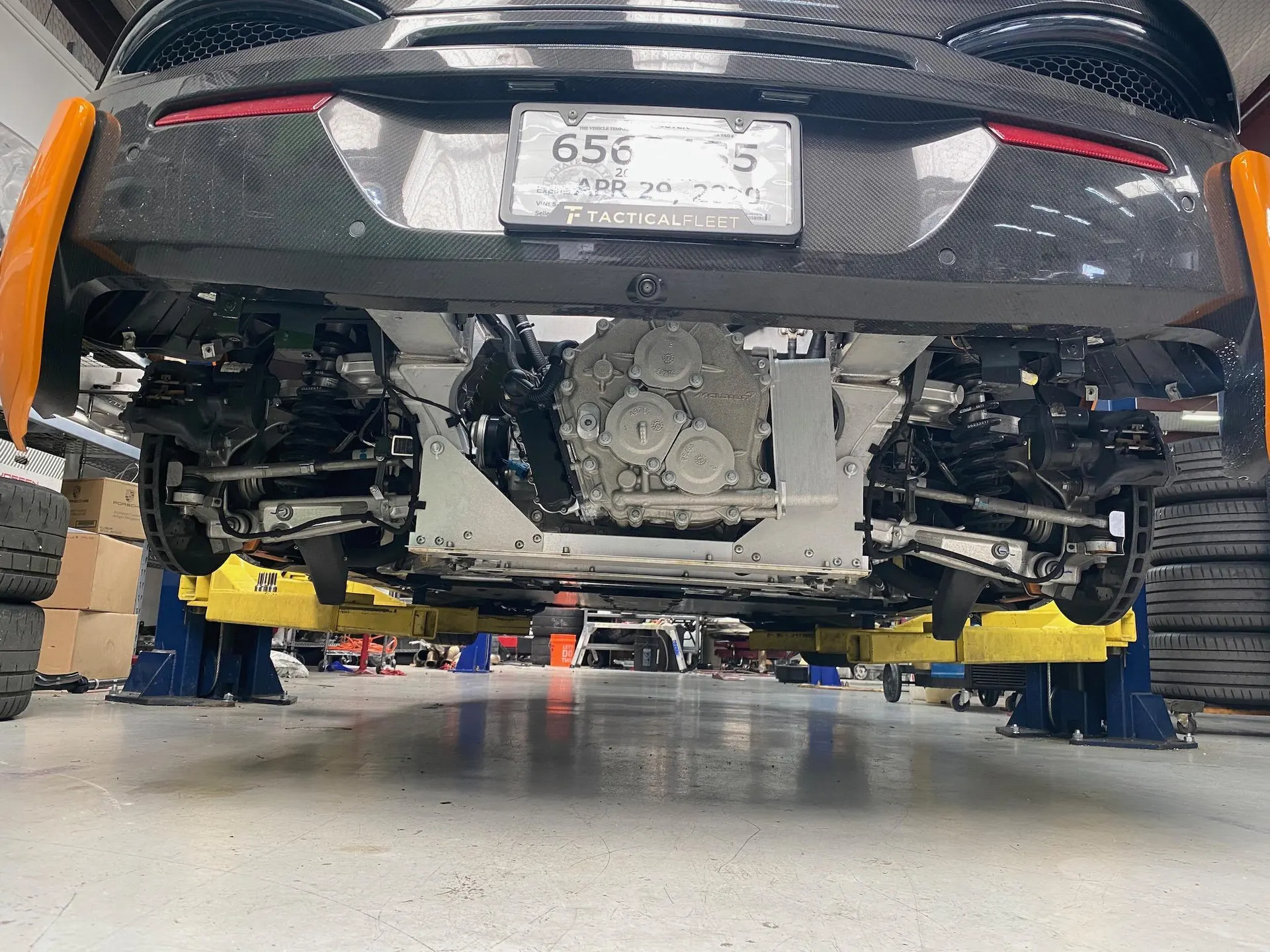 Underside view of a McLaren on a lift in a performance shop, showcasing the rear suspension, brakes, and drivetrain.