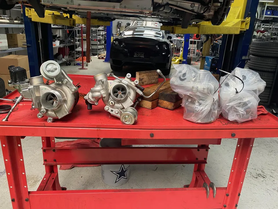 New and used turbochargers on a red workbench in a performance tuning shop with a car on a lift in the background.