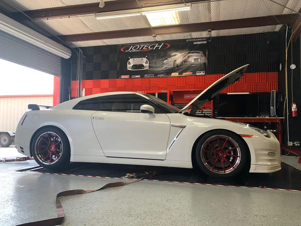A white Nissan GT-R with red wheels on a dyno at Jotech Motorsports tuning shop, hood open for tuning.