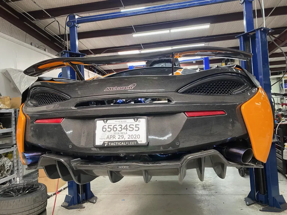 Rear view of a McLaren on a shop lift, featuring a large carbon fiber rear wing, diffuser, and aftermarket exhaust.