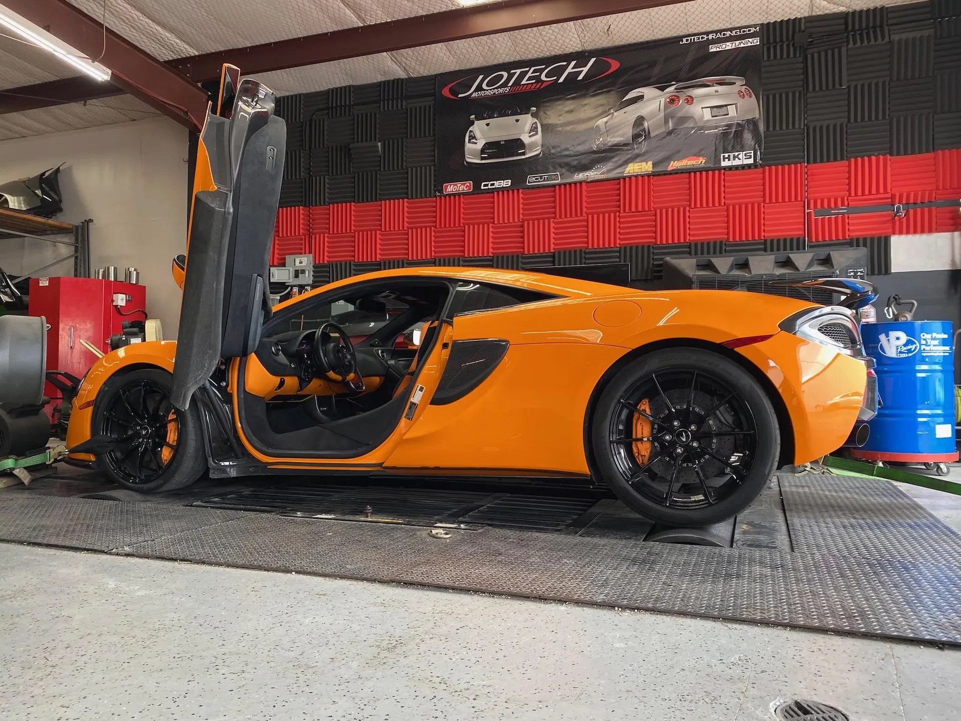 An orange McLaren supercar with its doors open, strapped to a dyno at Jotech Motorsports, ready for a performance test.