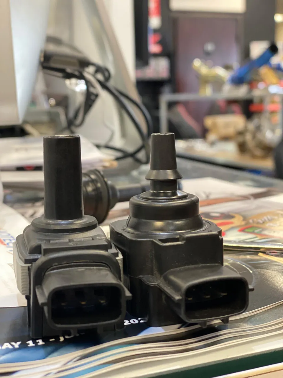 Two black ignition coils on a desk, likely for a performance vehicle, in a shop setting.